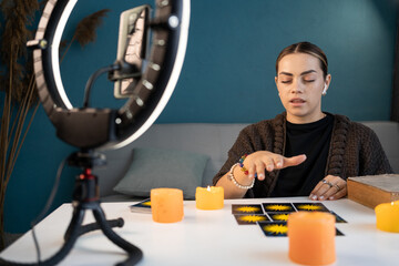 Female fortune teller read tarot cards online to predict fortune. Future reading with smartphone and ring lamp at home. Ritual candles burning.