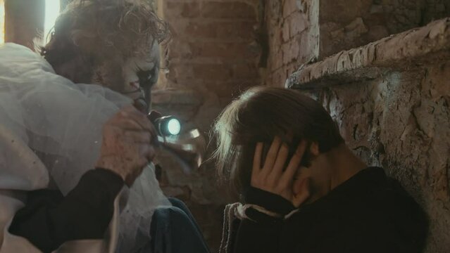 Creepy manic in clown costume scaring girl with sounds of horns while sitting on floor in abandoned place