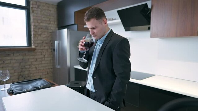 Side angle view thoughtful man in elegant suit smelling red wine in glass standing in kitchen at home indoors. Young Caucasian freelancer businessman with alcohol on weekend
