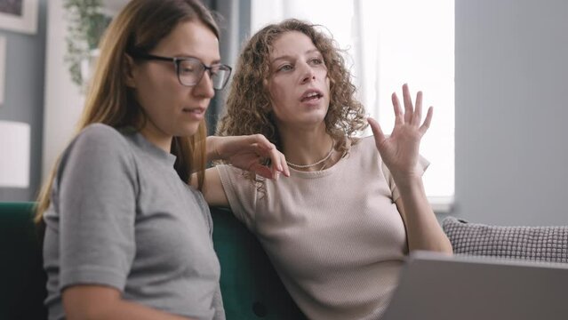 Two friends browsing on laptop on couch, curly woman explain friend in glasses what exactly need and together search for this on Internet