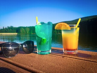 Two colorful cocktails and a pair of sunglasses placed on a wooden pontoon near the lake in summer