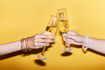 Vibrant close up image of two people clinking champagne glasses on yellow background in celebration