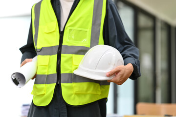Architect man in reflex vest holding hardhat and blueprints standing in office