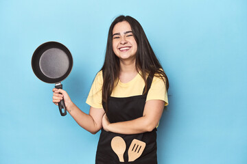 Young woman with pan on blue studio laughing and having fun.