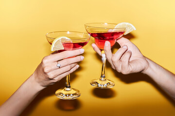 Close up of two hands clinking cocktails against vibrant yellow background in celebration, copy...