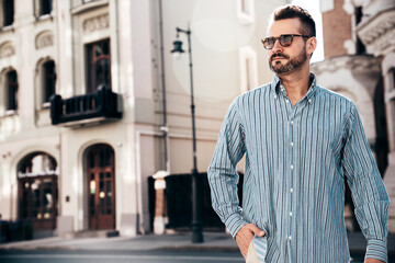 Obraz premium Portrait of handsome confident stylish hipster lambersexual model. Sexy modern man dressed in blue shirt and trousers. Fashion male posing on street background in Europe city at sunset. In sunglasses