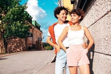 Naklejka premium Young smiling beautiful woman and her handsome boyfriend in casual summer clothes. Happy cheerful family. Female having fun. Couple posing in the street at sunny day. Near wall at sunset