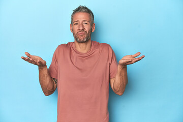 Middle-aged caucasian man on blue backdrop doubting and shrugging shoulders in questioning gesture.