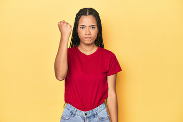 Young Indonesian woman on yellow studio backdrop showing fist to camera, aggressive facial expression.