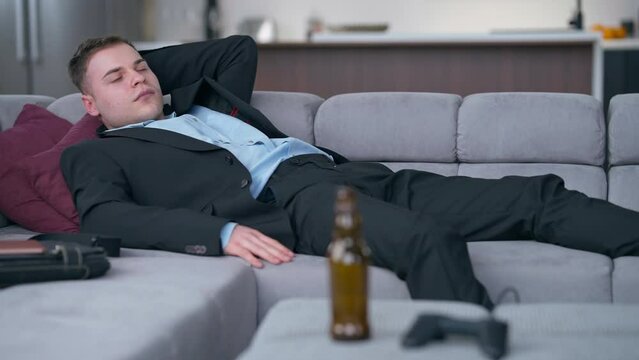 Portrait of exhausted young man in suit on couch with blurred beer bottle and game controller at front. Tired Caucasian freelancer on Friday at home in living room