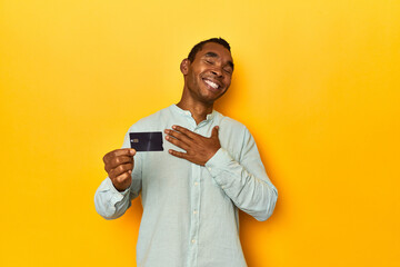 African American man with credit card, yellow studio, laughs out loudly keeping hand on chest.