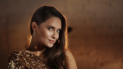 Closeup shot of beautiful brunette woman actress in golden dress standing against wall and posing smiling at the camera.