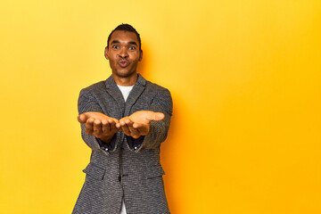 African American man in casual blazer, yellow studio, folding lips and holding palms to send air kiss.