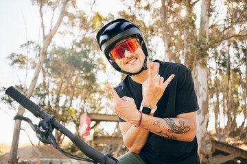Obraz premium Rock sign, bike and man with helmet in forest on off road cycling workout with happiness and glasses. Portrait, person and smile with hand gestures on bicycle for exercise, challenge and competition