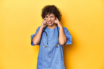 Curly-haired Caucasian woman nurse on yellow studio covering ears with hands.