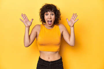 Curly-haired Caucasian woman in yellow top celebrating a victory or success, he is surprised and...
