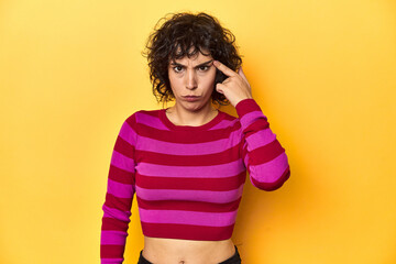 Caucasian curly-haired woman in pink striped-top pointing temple with finger, thinking, focused on...