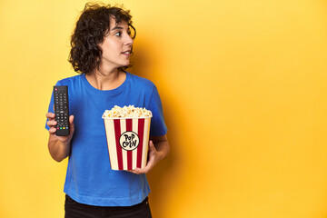 Woman with popcorn and TV remote, home cinema concept, yellow studio backdrop.