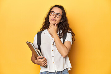 Student holding notebooks, glasses, backpack on, looking sideways with doubtful and skeptical expression.