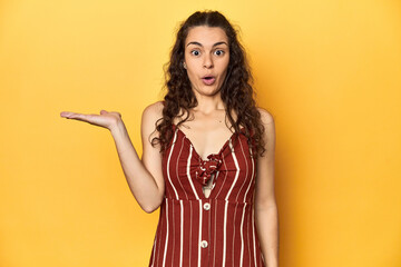 Young Caucasian woman, yellow studio background, impressed holding copy space on palm.
