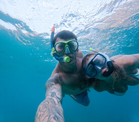 Beautiful and cute couple of millennials young people snorkeling and diving together looking underwater for fishes and coral having fun and enjoying. Summer time swimming in the sea blue ocean.
