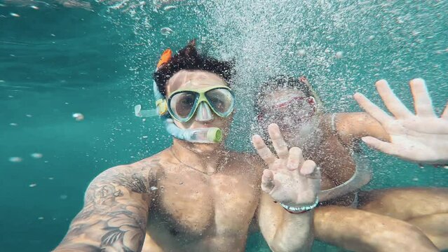 Beautiful and cute couple of millennials young people snorkeling and diving together looking underwater for fishes and coral having fun and enjoying. Two beautiful people waving underwater at camera
