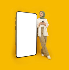 New mobile application, full body young smiling caucasian muslim girl in hijab using smartphone offering new mobile phone application. Empty blank screen huge phone mock up. Yellow background.