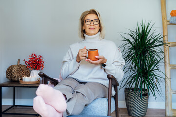 Middle aged woman relaxing with cup of hot drink in scandy style hygge interior home with fall mood...