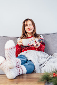 Relaxed woman in plaid and warm socks drinking hot tea with remote controller on sofa at home while watching movie, TV with christmas decoration atmosphere. Cozy and comfortable winter. Vertical