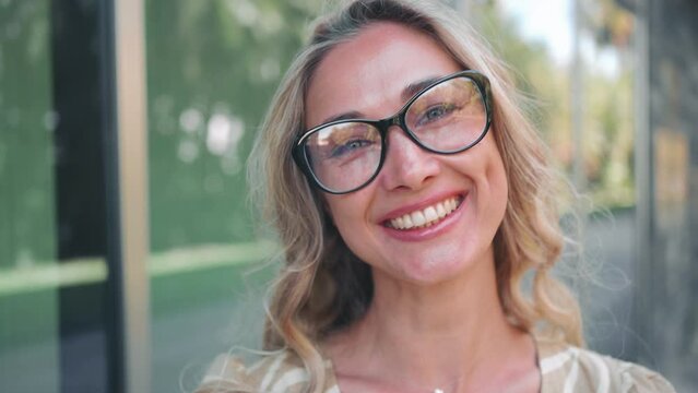 Pretty smiling joyfully female with curly hair, dressed casually, looking with satisfaction at camera, being happy. Outdoor portrait of good-looking beautiful woman in glasses standing near office