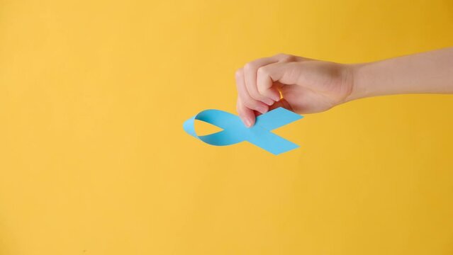 Vertical video of female hand hold blue ribbon, isolated over yellow studio background wall with copy space. Healthcare, International men, Father and World cancer day concept. Symbol for support men