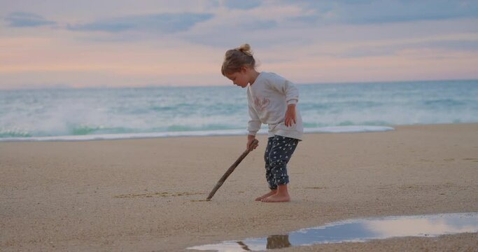 Little beautiful girl draws with a stick on the sand on the ocean shore at sunset