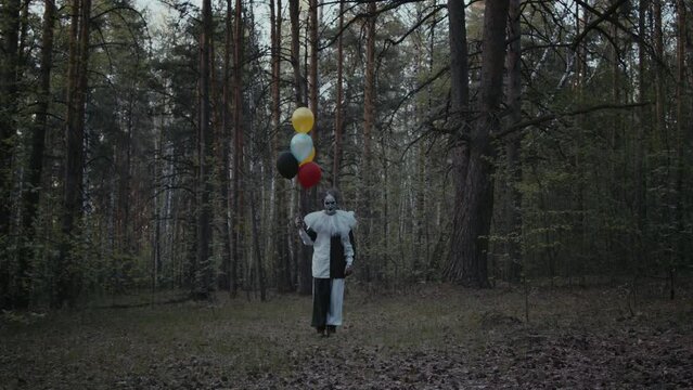 Wide shot of clown in black and white clothes holding colorful balloons in dark forest, looking at camera