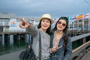 cheerful asian taiwanese woman showing friend landmark in distance with pointing gesture while using navigation app on phone during their trip at santa Barbara harbor in california