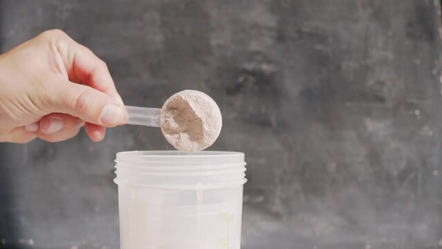 Male hand puts into a shaker portion of chocolate whey protein powder from measuring scoop on dark background, slow motion, close-up view