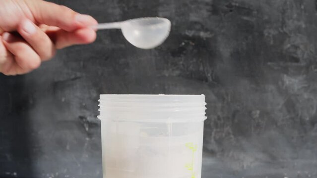Male hand puts into a shaker portion of chocolate whey protein powder from measuring scoop on dark background, close-up view