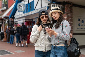 two asian japanese girl tourists searching right direction to go with navigation app on cellphone on the street during their spring travel in solvang Danish village ca usa - obrazy, fototapety, plakaty