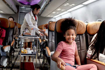 Happy African girl child, kid passenger sitting in comfortable seat inside airplane during female air hostess pushing cart on aisle to serve food and drink beverage, airline service and transportation