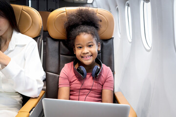 Happy curly hair African girl child passenger with headphones using laptop computer, sitting in seat inside airplane, cheerful kid enjoys traveling by airline transportation, holiday adventure trip