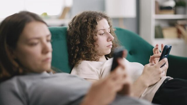 Two bored women on couch, looking at smartphones and ignoring each other, feeling lonely at home during the day