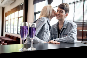 Cocktail drink glasses with blurred background of cute couple kissing while sitting by table in bar at airport lounge, having drinks during waiting flight, happy lover enjoy romantic date in club.