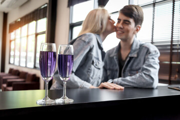 Cocktail drink glasses with blurred background of cute couple kissing while sitting by table in bar at airport lounge, having drinks during waiting flight, happy lover enjoy romantic date in club.