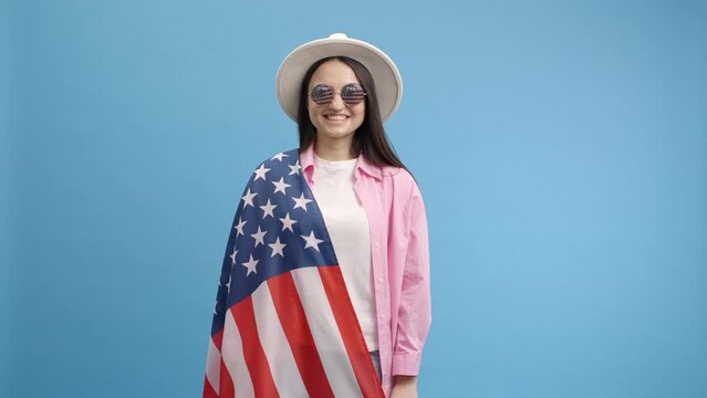 Trendy cheerful positive girl in white hat and glasses holds an American USA flag over his shoulder, celebrating. Independence day. Young pretty adult woman. Indoor studio shot on blue background