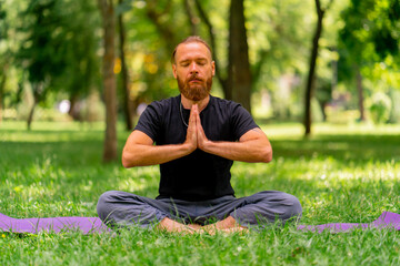 Naklejka na ściany i meble portrait handsome bearded man meditating in the park sitting in the lotus position practicing yoga zen healthy lifestyle calm meditation