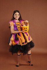 happy girl in colorful Halloween attire holding bucket with candies  and standing on brown backdrop