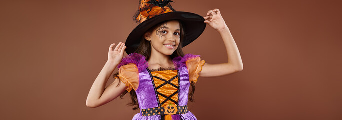 happy girl in Halloween costume and pointed hat posing on brown background, little witch banner