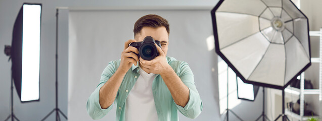 Male photographer working in studio. Front view of young man standing in studio with grey background and lighting equipment, holding camera, and taking photo. Professional photography concept. Banner
