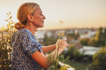 Beautiful woman enjoys drinking coffee while standing on her balcony at sunset. - obrazy, fototapety, plakaty