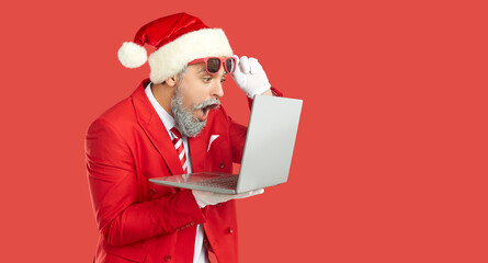 Man in Santa suit opens mouth and looks at laptop screen with surprised face expression impressed...