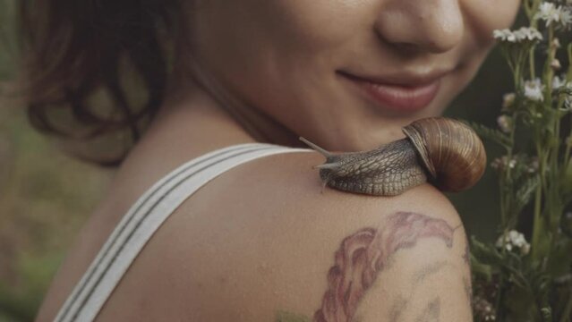 Close-up of snail slowly crawling along shoulder of young girl. Woman holding bouquet of wild flowers or daisies in her hands. Snail leaves muscus on skin. Concept of nature, cosmetology, spa, beauty.
